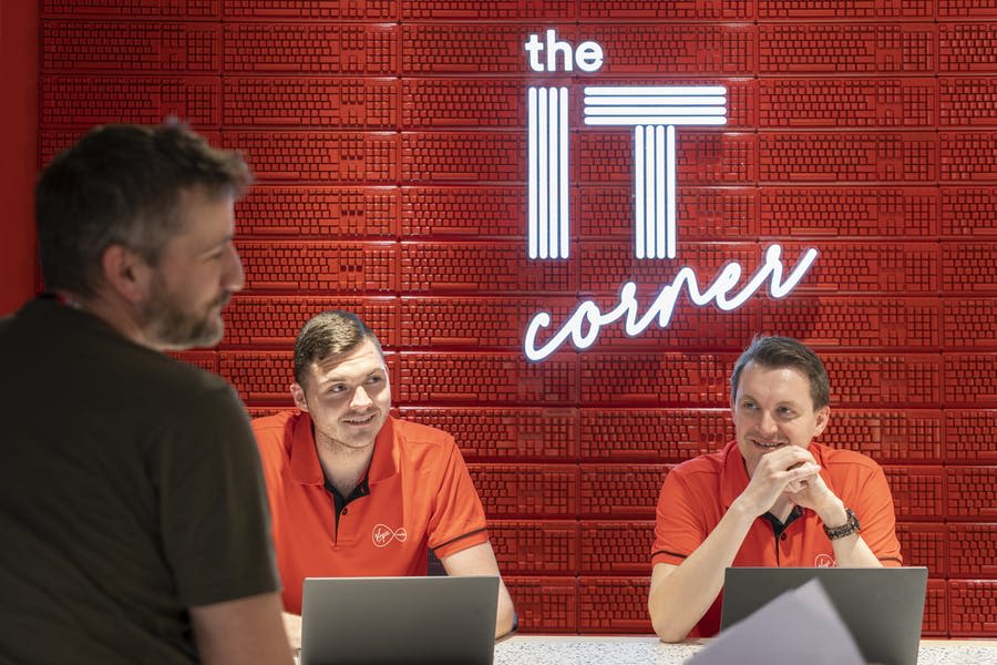 Two men with laptops sit in front of a red wall with neon lights spelling out The IT Corner on it. Another man is approaching them.