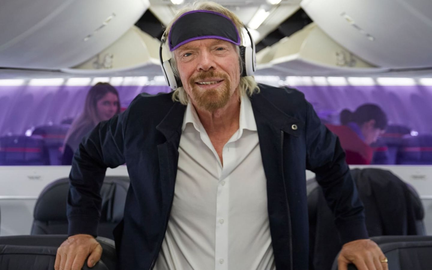 Richard Branson standing in the aisle of a plane wearing headphones
