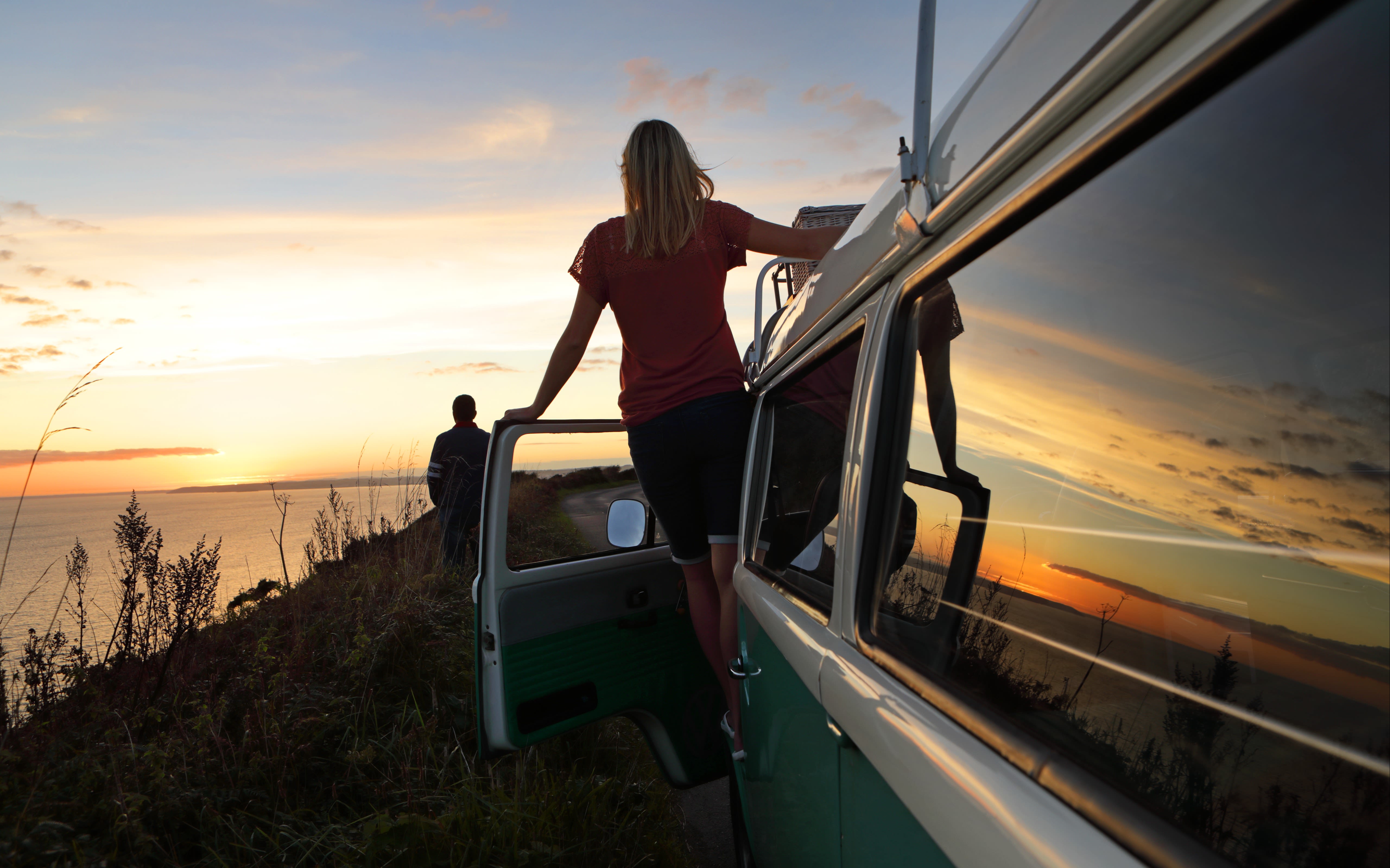 Image of a woman standing out the side of a camper van, and a man further ahead, both looking at the sunset by the coast in Cornwall. 
