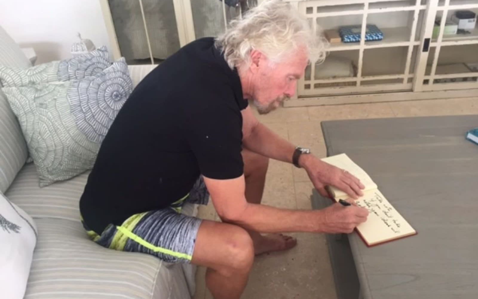 Richard Branson sitting on a sofa and writing in a notebook 