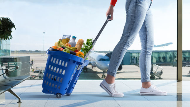 An image of a person wheeling a blue, filled Tesco shopping basket through an airport terminal. 