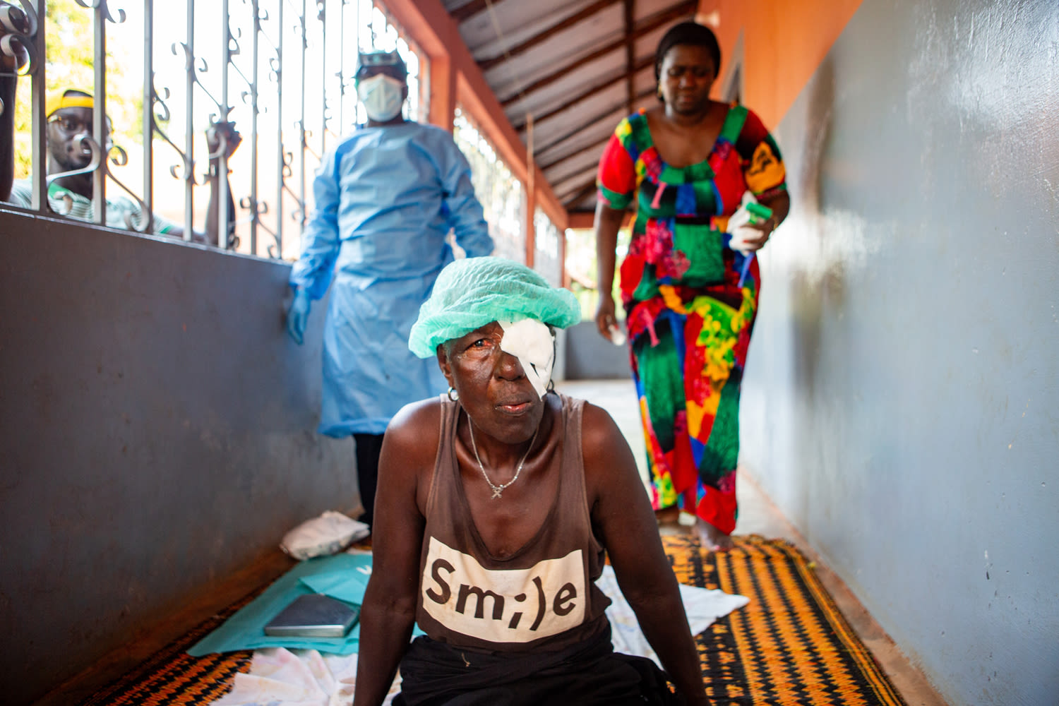 Nene sits up following trachoma surgery on the porch of her home while Forma,trachoma surgeon and his assistant Dionesa clean up from the surgery