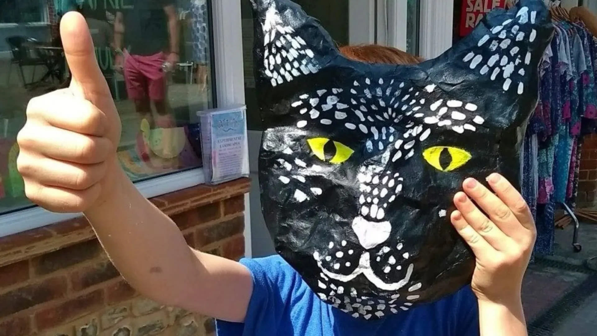 A child giving a thumbs up while holding a cat mask they have made up to their face