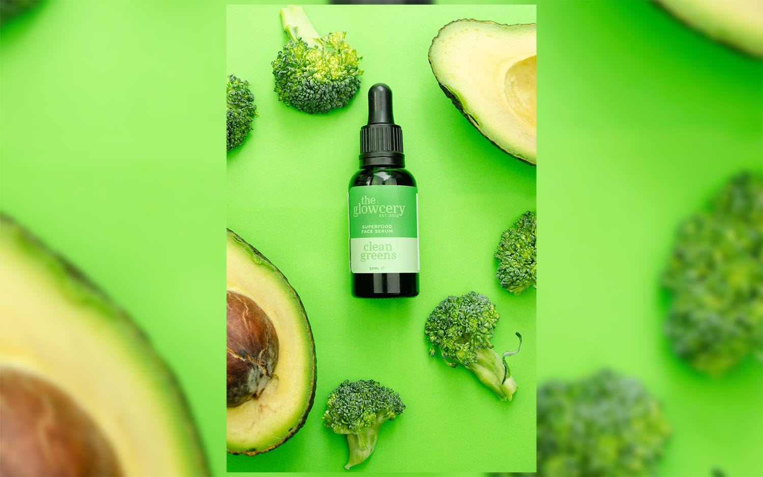 A bottle of The Glowcery's superfood serum on a green background surrounded by broccoli and avocado
