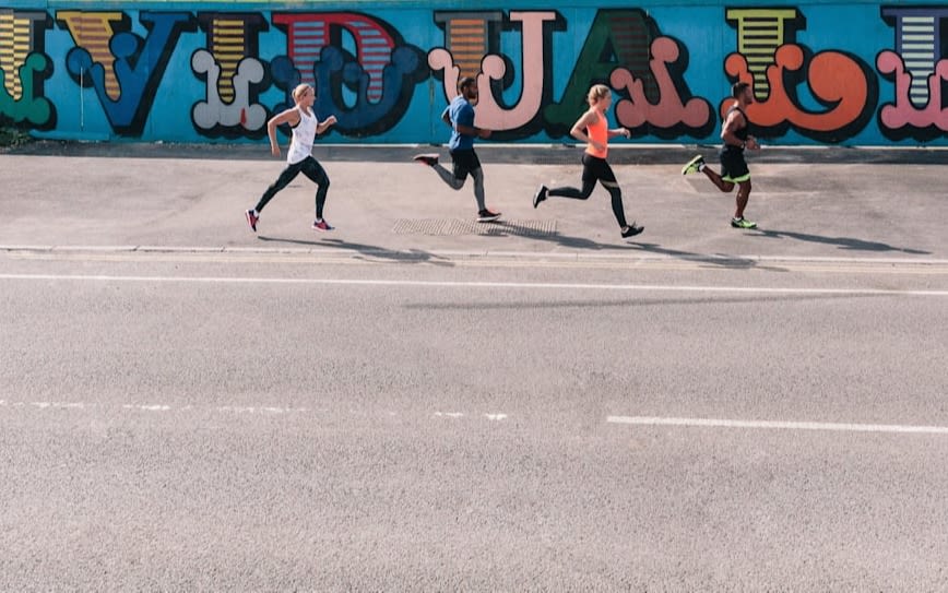 2 men and 2 women running on a pavement to their left is a graffiti wall spelling individuality and to their right is a road.