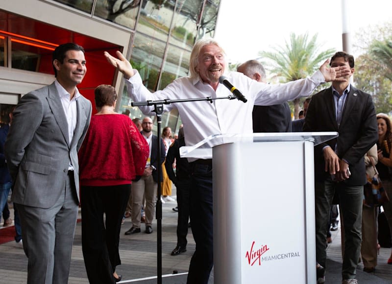 Richard Branson speaking into a microphone behind a podium, with his arms out to the side