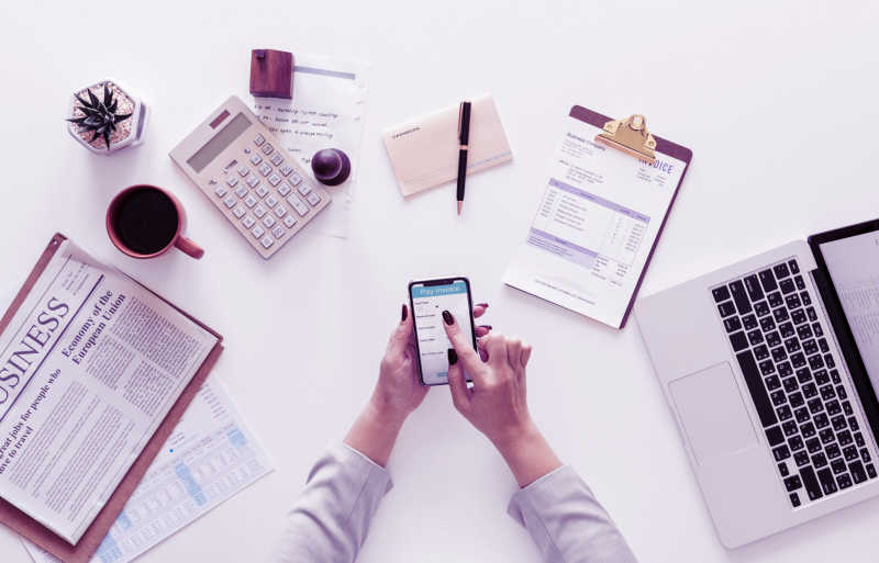 Illustration of top view of  woman's hands using a mobile phone surrounded by laptop, clipboard, cashbook, pen, calculator, newspaper, potted plant, and mug