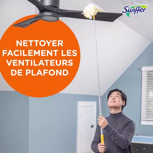 Une personne utilise un Swiffer pour nettoyer un ventilateur de plafond dans une pièce lumineuse aux murs bleu clair et aux bordures blanches.