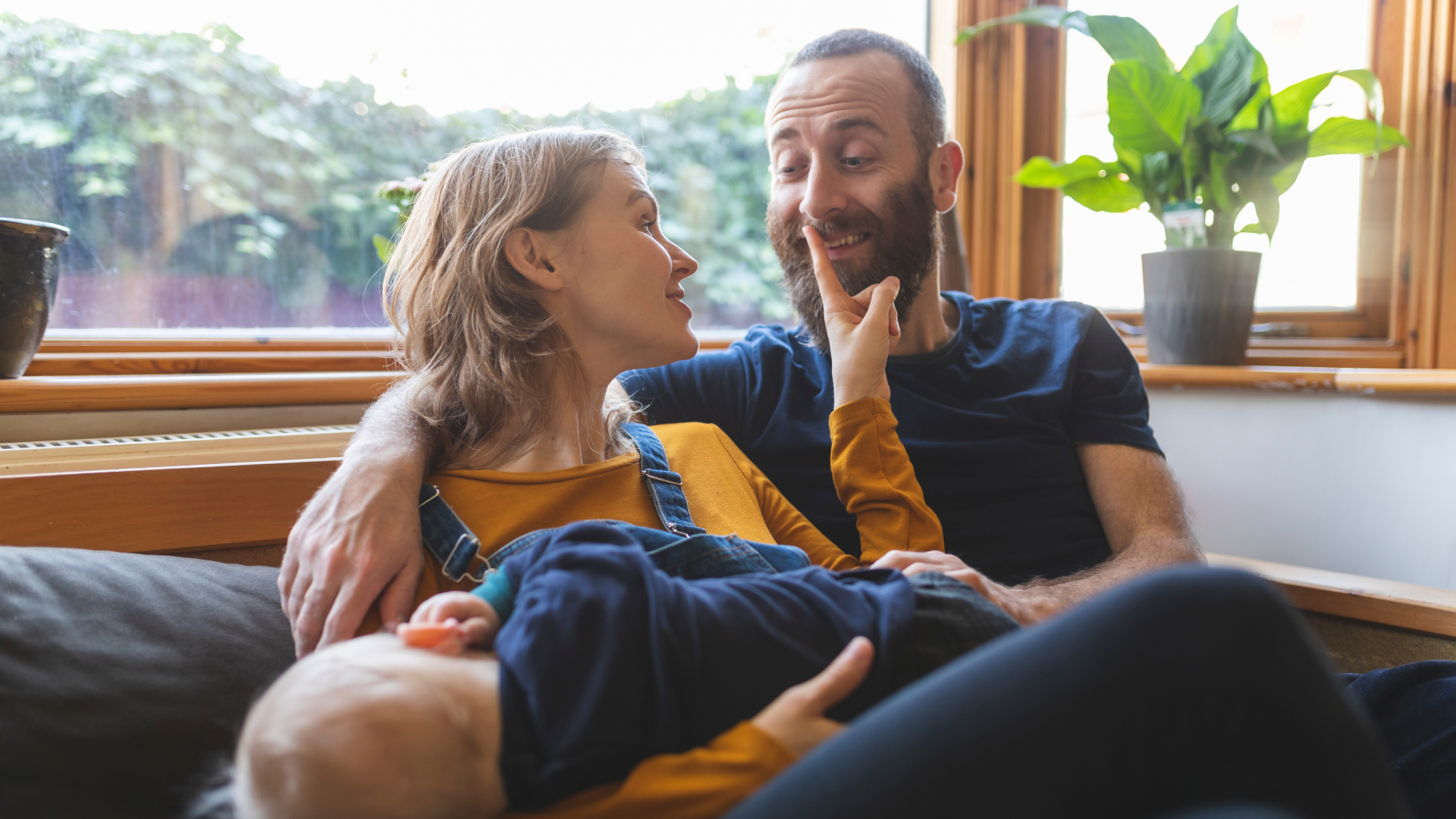 A woman and man sitting on a sofa, smiling and playing with their baby