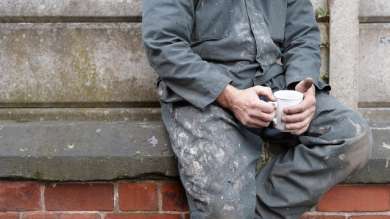A tradesman holding a mug whilst sat on a brick wall.