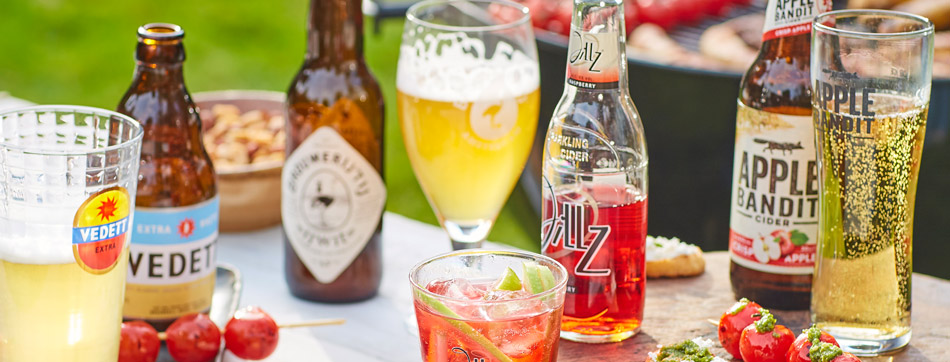 Diverse zomerse drankjes in flessen en glazen, waaronder bier, cider en een cocktail. Merken als Vedett, Apple Bandit en Jillz zijn zichtbaar.