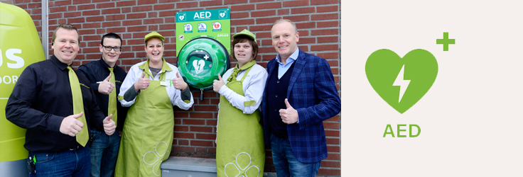 Groepsfoto met vijf PLUS-medewerkers voor een muur, bij een AED-apparaat.