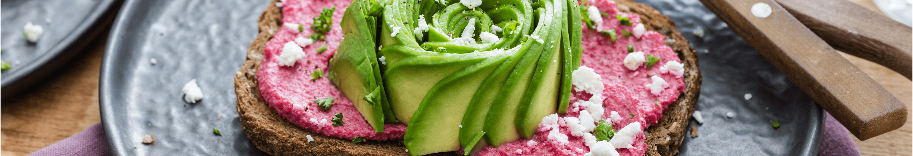 Een snee toast met bietenhummus, afgewerkt met een avocado-roos, witte kaas en groene kruiden.