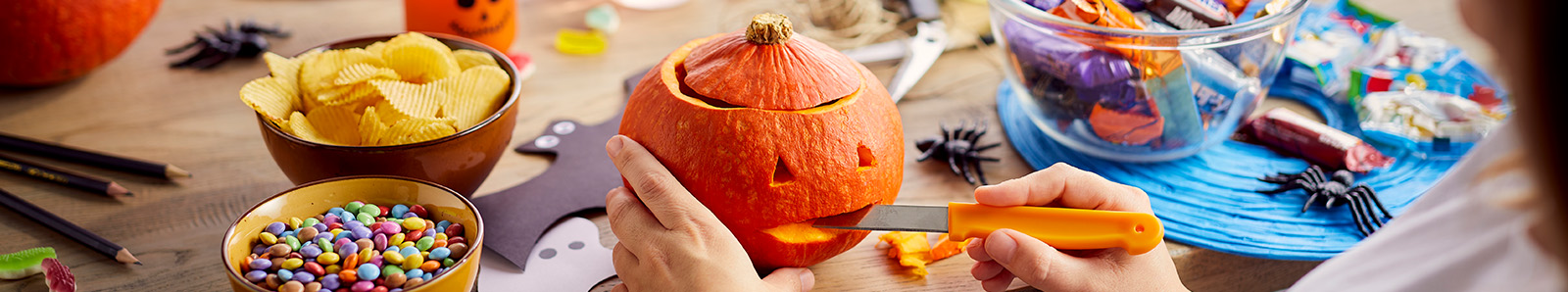 Persoon snijdt een pompoen met een oranje mes aan een tafel met Halloweenversiering, chips en snoep.