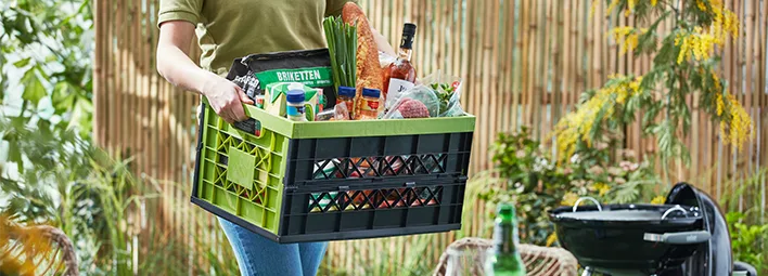 PLUS-bezorger draagt een PLUS-boodschappenkrat gevuld met producten als een stokbrood, fles drank, bosui, briketten, mayonaise.