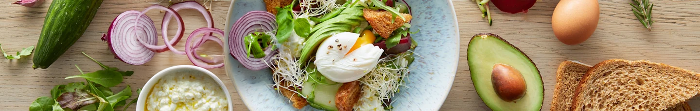 Vers ontbijt met gepocheerd ei op salade, omringd door avocado, komkommer, rode ui, volkorenbrood, ei en een kom cottage cheese of yoghurt.