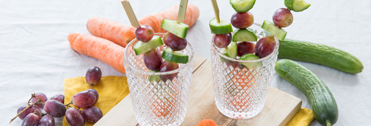 Gezonde snackspiesjes met druiven en komkommer in glazen, met wortels en komkommer op de achtergrond.