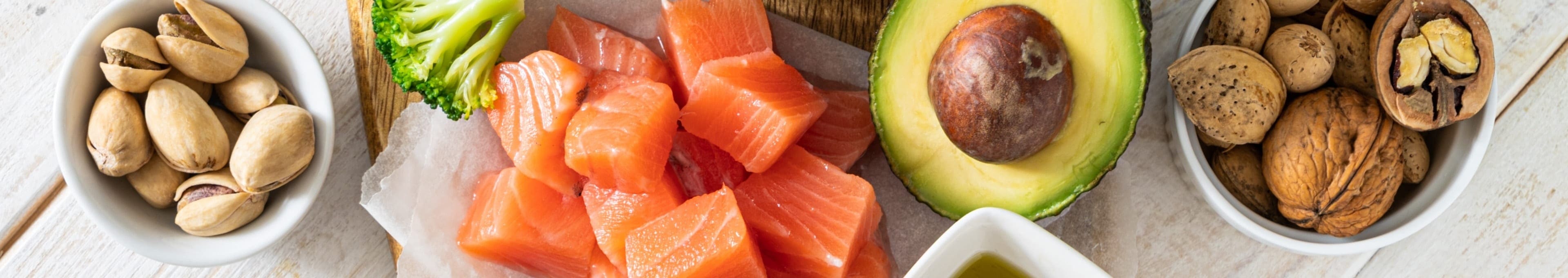 Een selectie van gezonde voedingsmiddelen, waaronder zalmblokjes, broccoli, avocado, pistachenoten, gemengde noten en een schaaltje olijfolie.