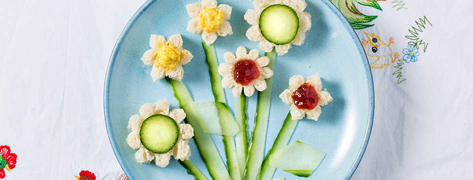 Ontbijt op een blauw bord: bloemen gemaakt van brood, komkommer en jam, met komkommerschijfjes als stelen en bladeren.