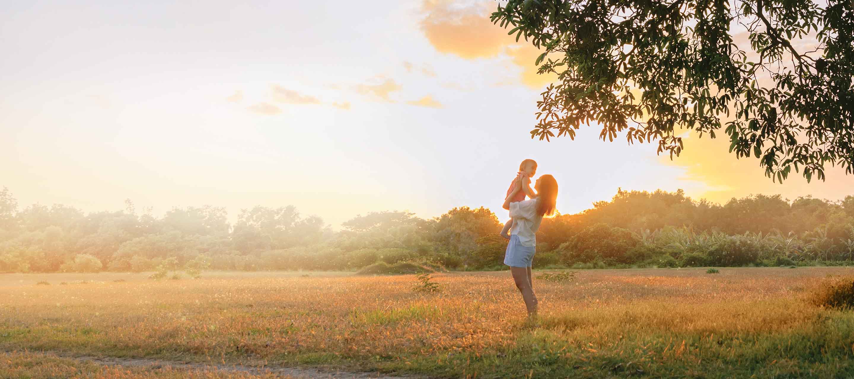 A mom raises her little girl in the air, sharing a tender moment in a grassy field during the summer at dusk.