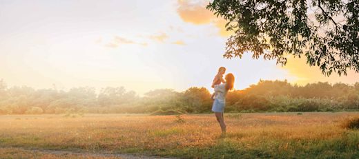 A mom raises her little girl in the air, sharing a tender moment in a grassy field during the summer at dusk.