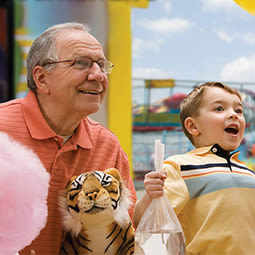 A picture of an old man holding a stuffed tiger and pink cotton candy standing next to a happy child in a yellow shirt holding a goldfish with the backdrop of an amusement park.