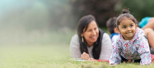 A young family of four is sitting on a blanket on the grass watching their baby daughter crawl.
