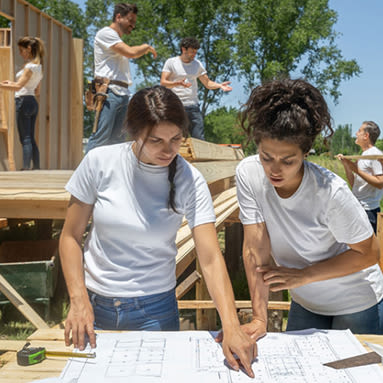 Two women review construction site plans while volunteers help build a house.