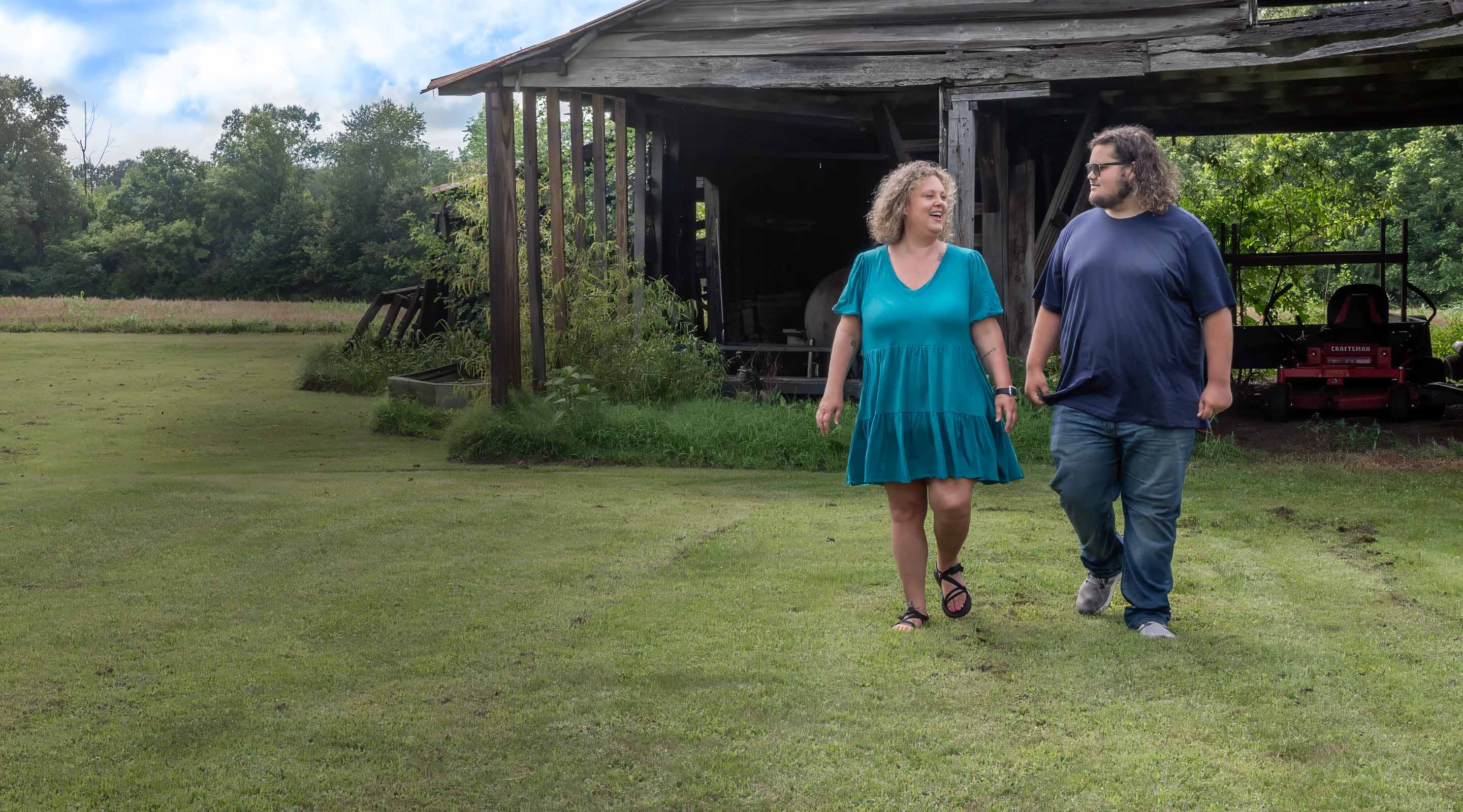 A mother and her son strolls through the grass near a weathered barn shelter.