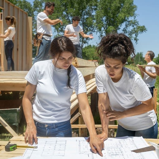 Two women referencing at blueprints, while others in the background work on building the foundation  and walls of the house