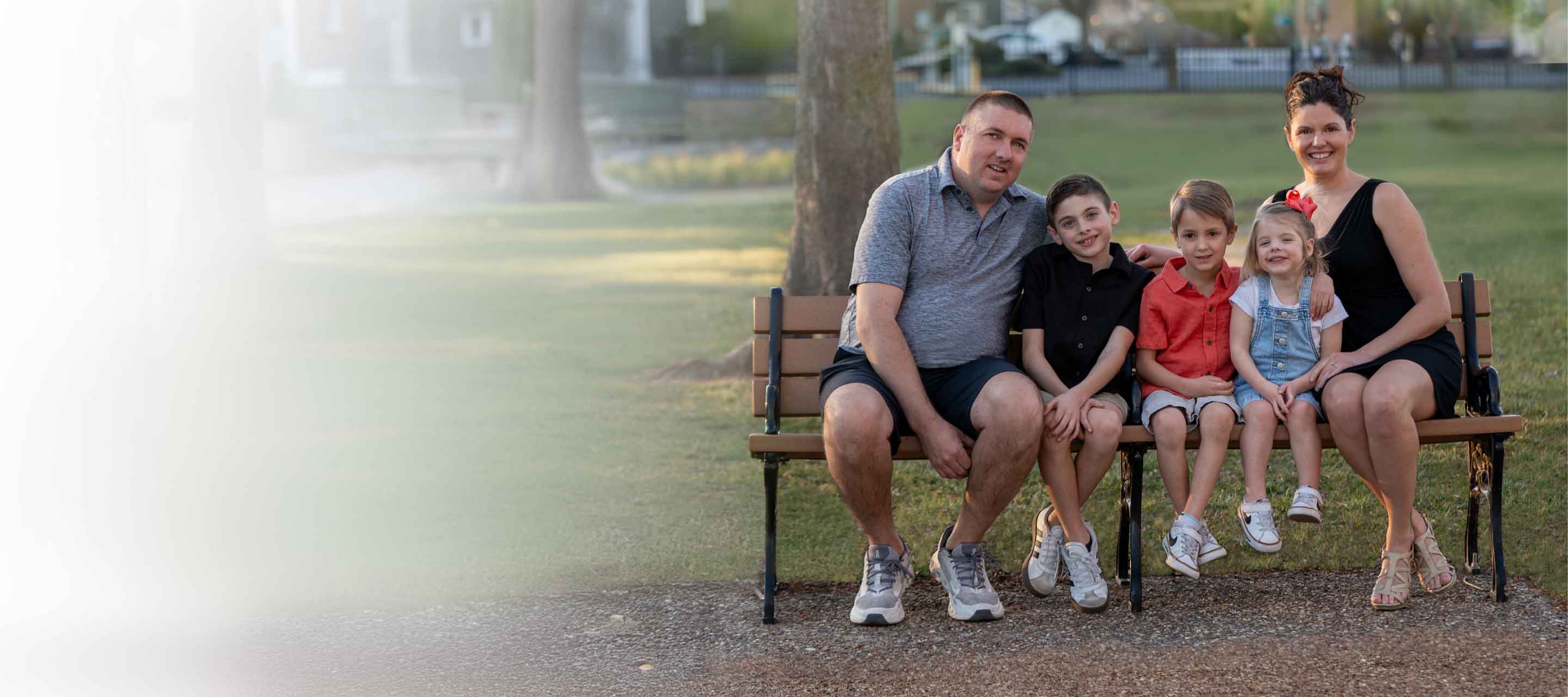 A family of five sitting on a park bench, including two parents and three children, smiling at the camera outdoors.