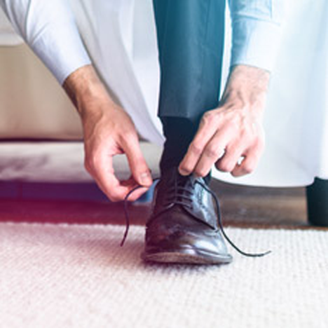 A close up of a man in blue slacks and a white button up bent over tying the laces of his derby dress shoe.
