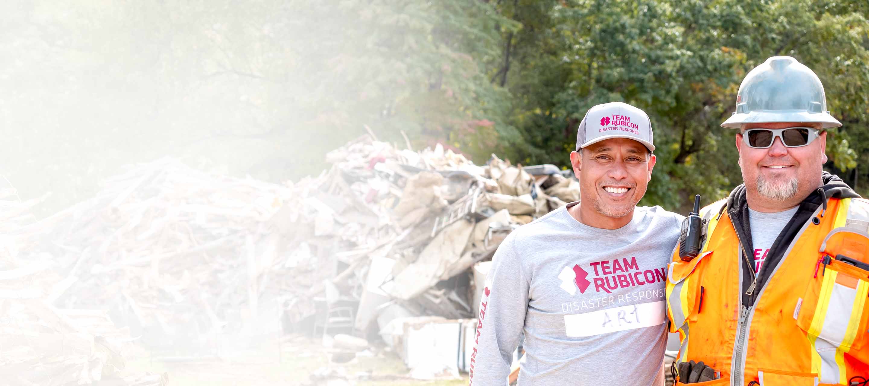 Two smiling Team Rubicon disaster response workers at a debris site, one in gray shirt and cap, one in orange safety vest.