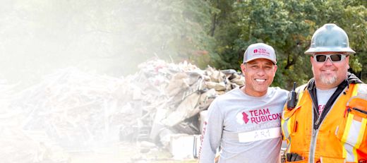 Two smiling Team Rubicon disaster response workers at a debris site, one in gray shirt and cap, one in orange safety vest.