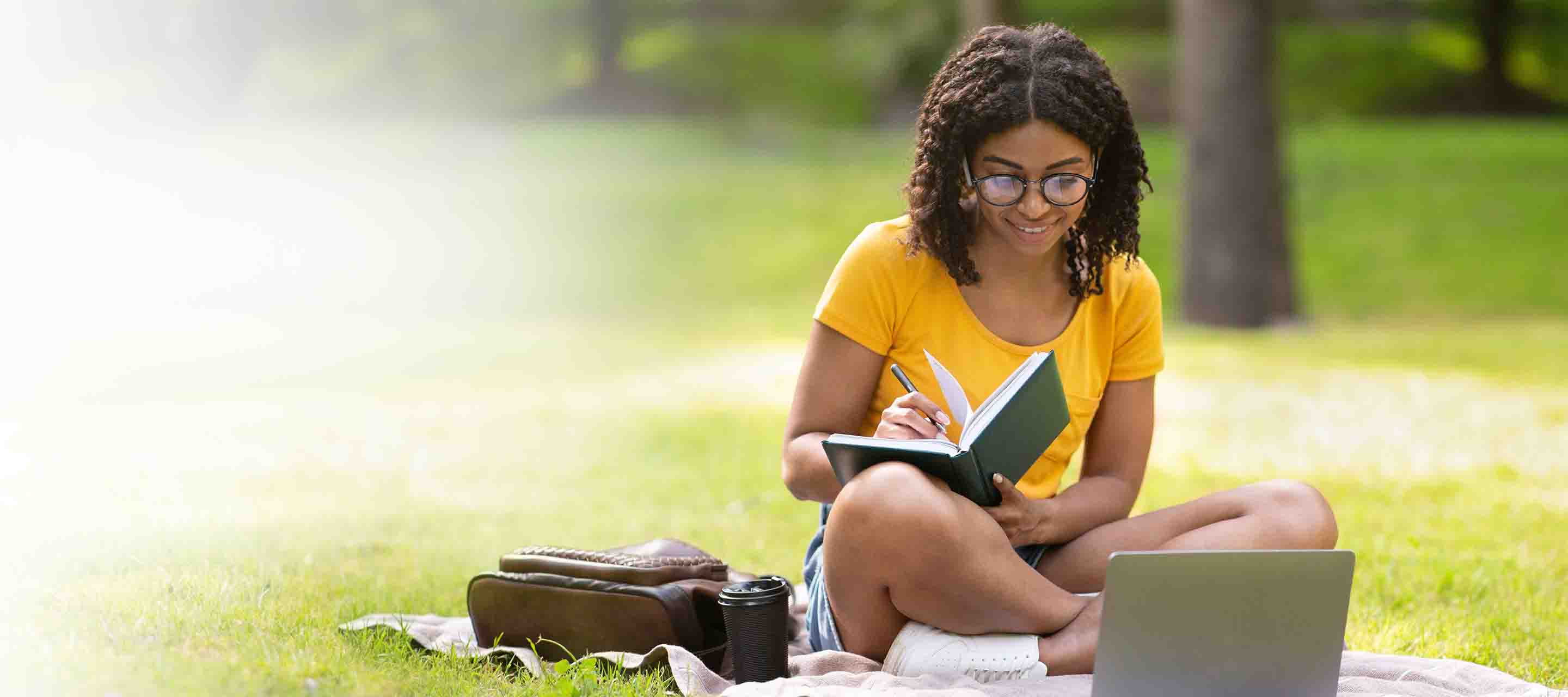 Young adult female is taking notes while sitting in a park watching her laptop.