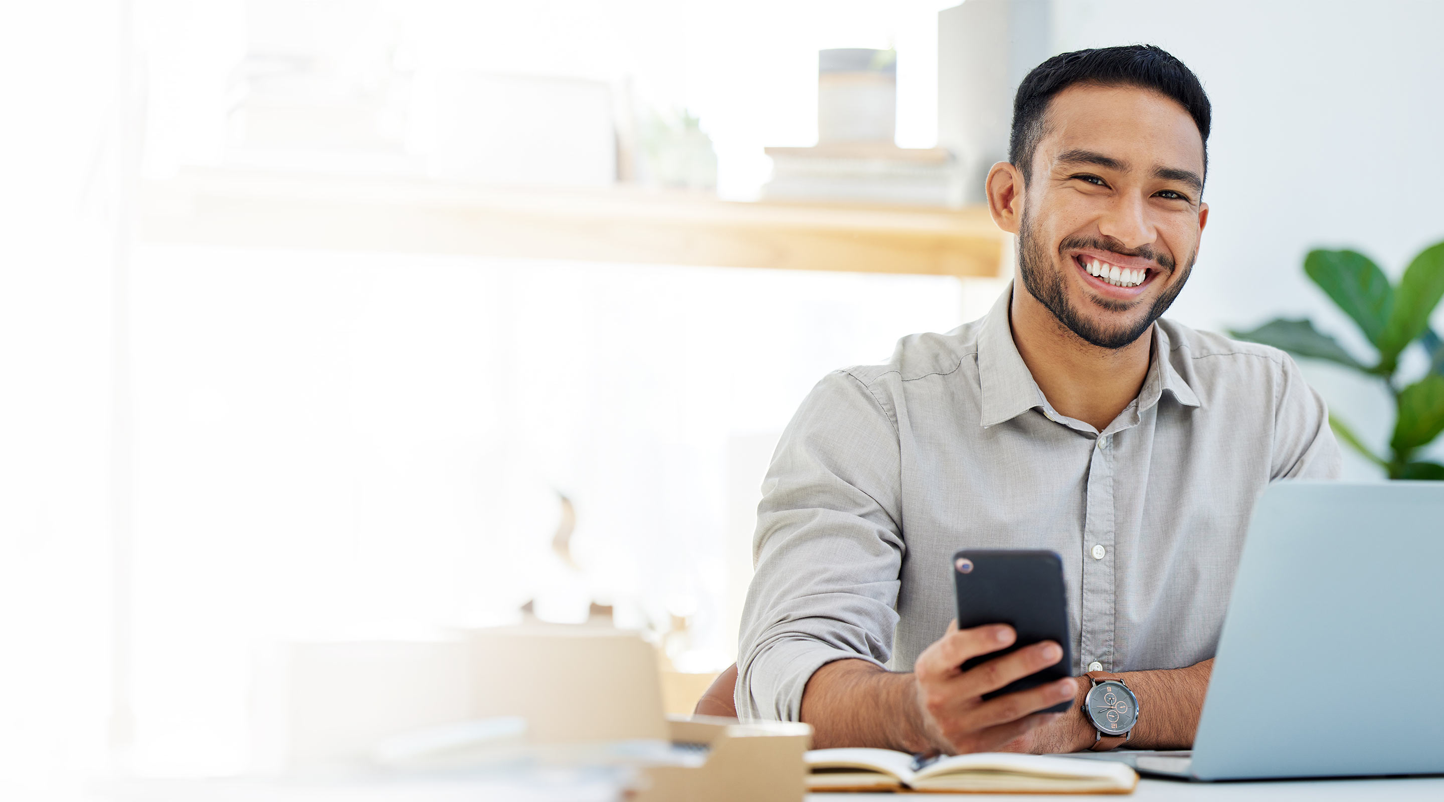 A smiling Asian man works at his computer and checks his phone