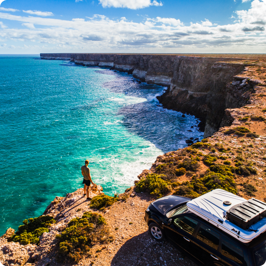 Traveler on a coastal cliff above turquoise ocean next to a camper SUV, a road trip booked with Zip buy now, pay later