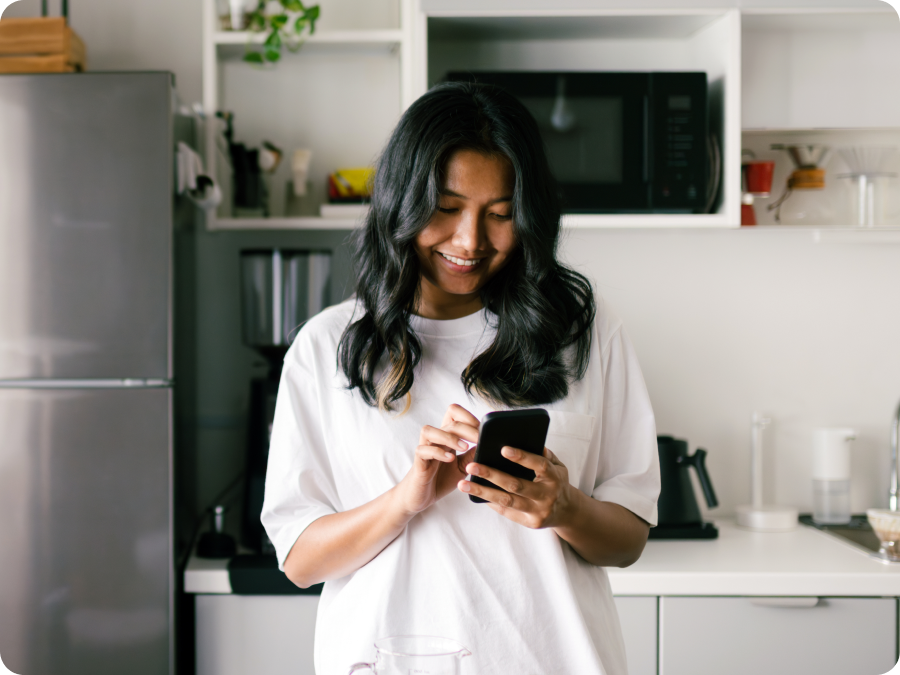 Woman in a white T-shirt using her smartphone in a modern kitchen to split a bill payment with Zip buy now, pay later