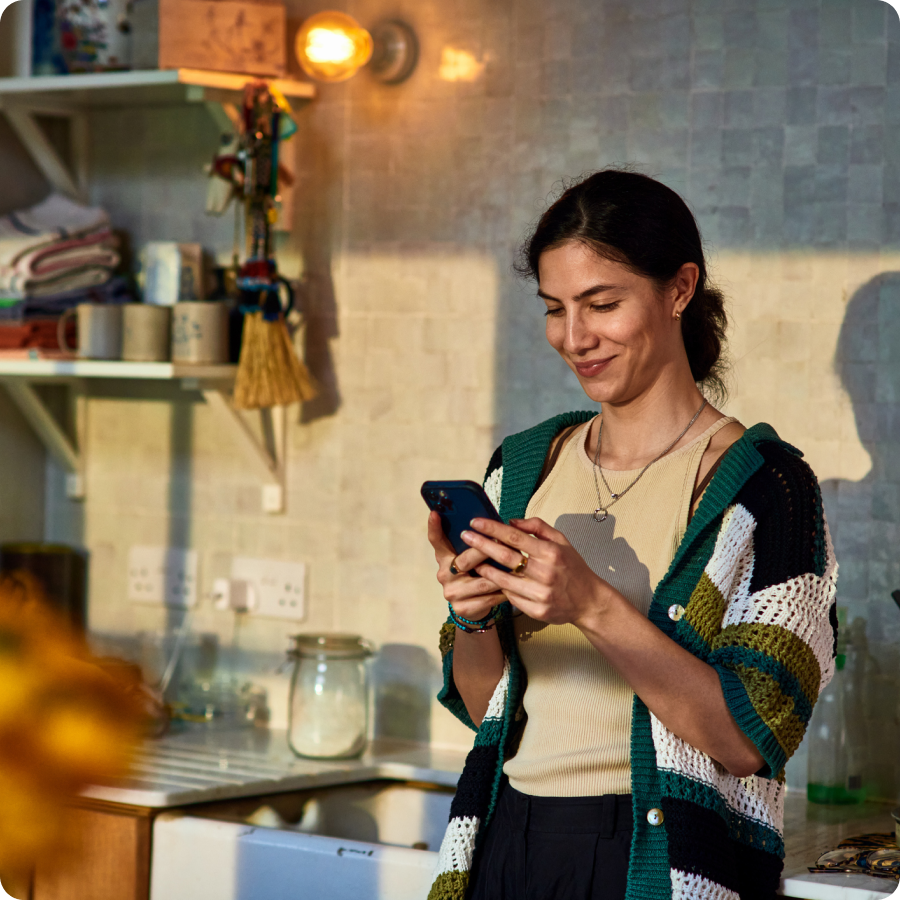 Woman in a cardigan smiling at her phone in a sunlit kitchen, paying bills in installments with Zip buy now, pay later