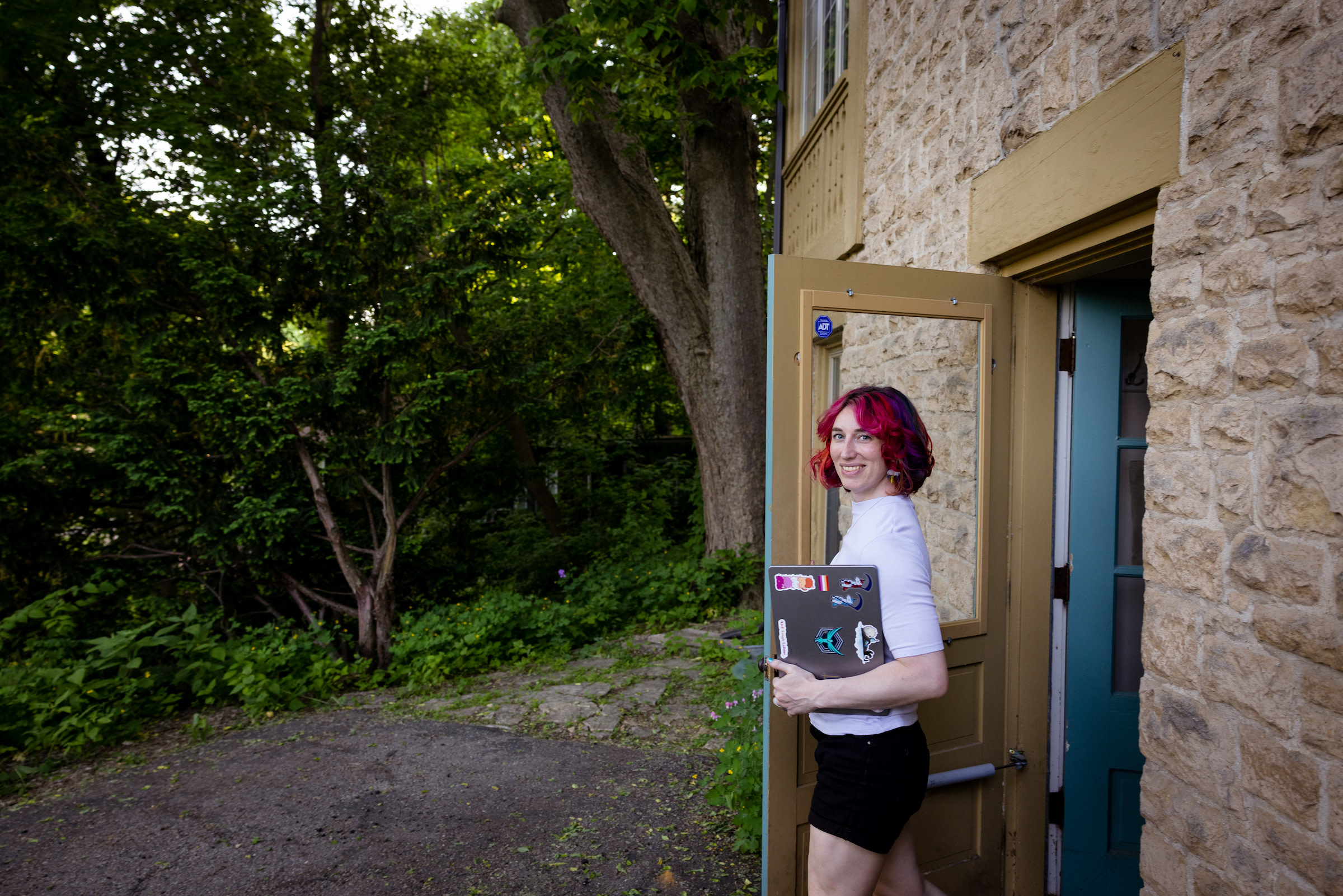Kyler stands outside at an open door, holding her computer and smiling.