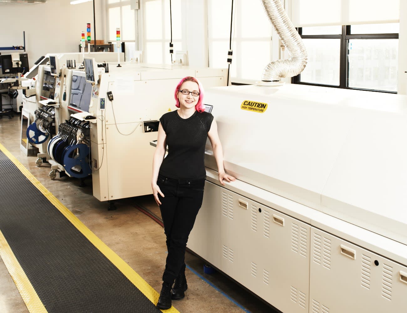 Photo of Limor Fried standing next to PCB production machinery and smiling.