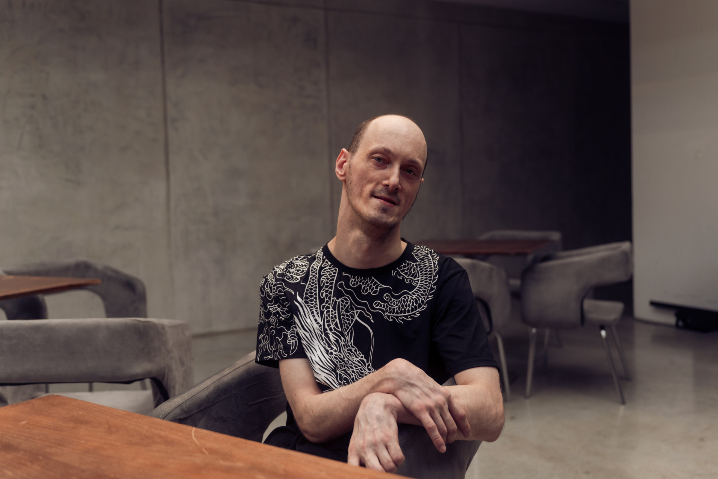 Anton sits in a gray chair with his arms crossed at a wooden table in a concrete building.