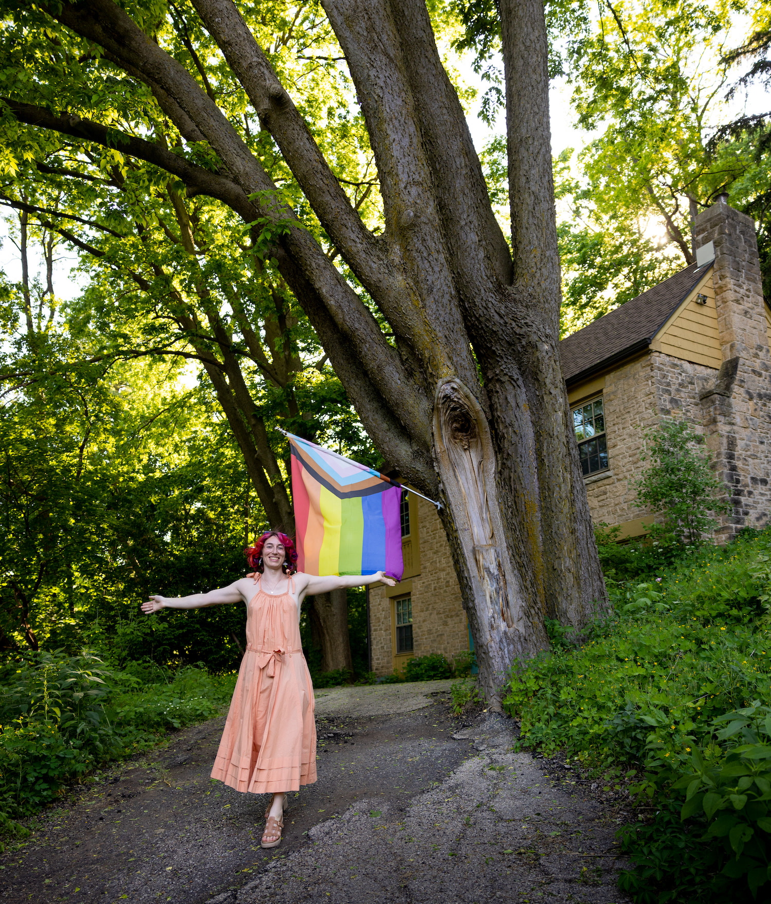 Kyler stands in front of her house, smiling with arms outstretched in front of a Pride flag.