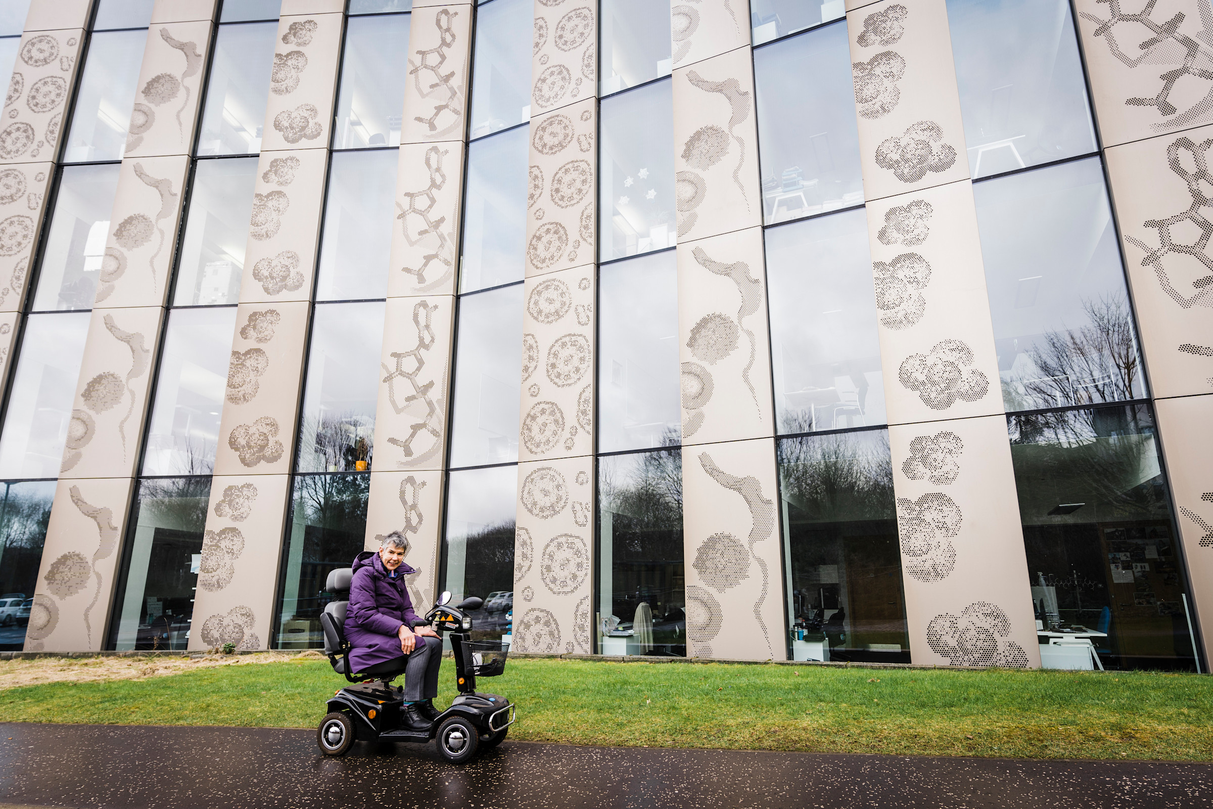 In a purple winter coat, Annalu sits in her motorized bicycle in front of a large building on the University of Dundee campus.