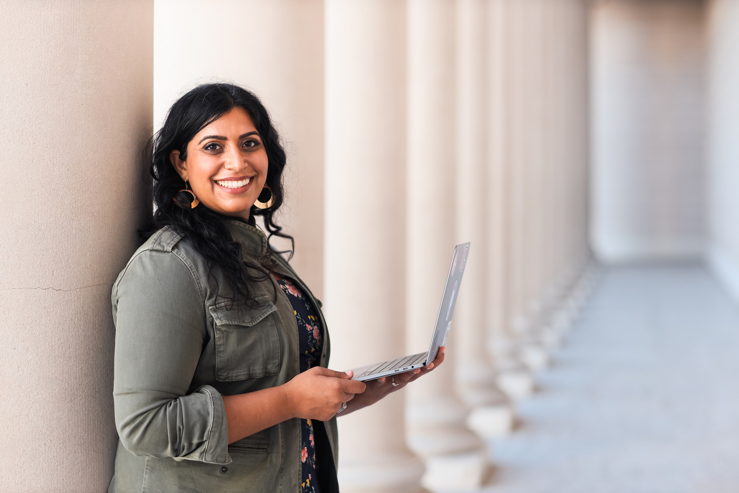 Author Neha smiling in a column-lined hallway with her laptop