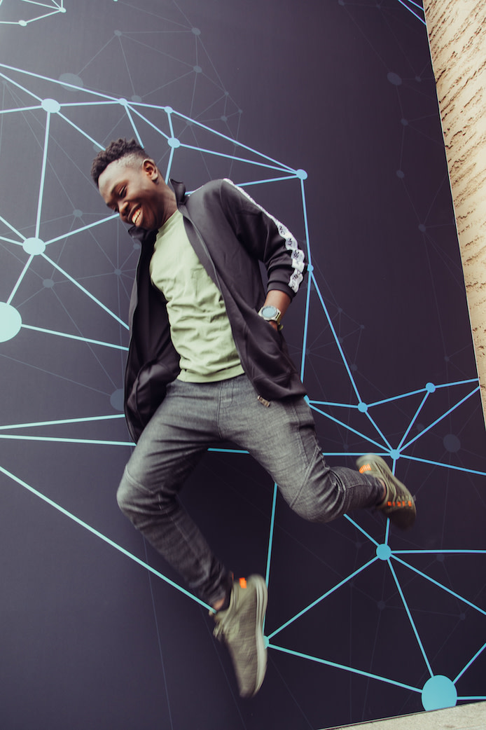 Samson Goddy is pictured jumping in mid-air, wearing earth tones and a smile.