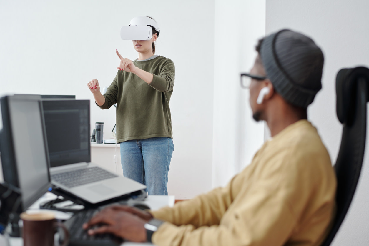 One person working on laptop while watching another person in a virtual reality headset