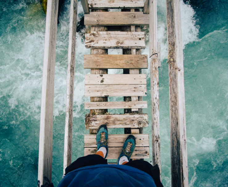 Person standing on a rickety wooden bridge over rushing water.