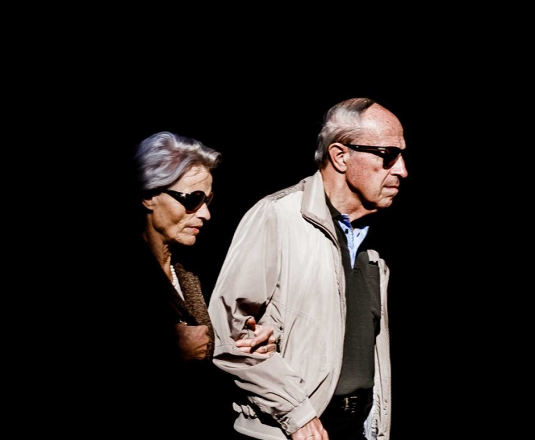 A man and woman, both wearing dark glasses, walking alongside each other, arm in arm.