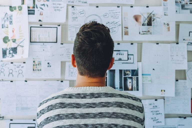 A person looks up at a white wall covered in different images, diagrams, and notes that have been drawn and printed out on paper.